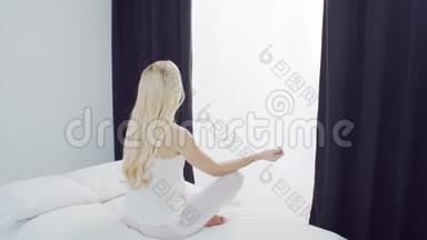 年轻女子在床上打坐.. 漂亮的金发女孩做瑜伽运动。 早上在卧室里，白天